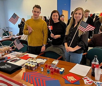 Student, studentka i Wykładowczyni stoją z flagami USA i Kanady uśmiechając się. Przed nimi biurko z materiałami promocyjnymi.