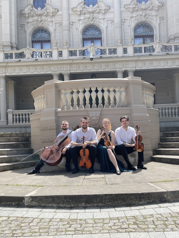 A woman and 3 men – members of the Vivenda String Quartet with string instruments sitting in front of the building