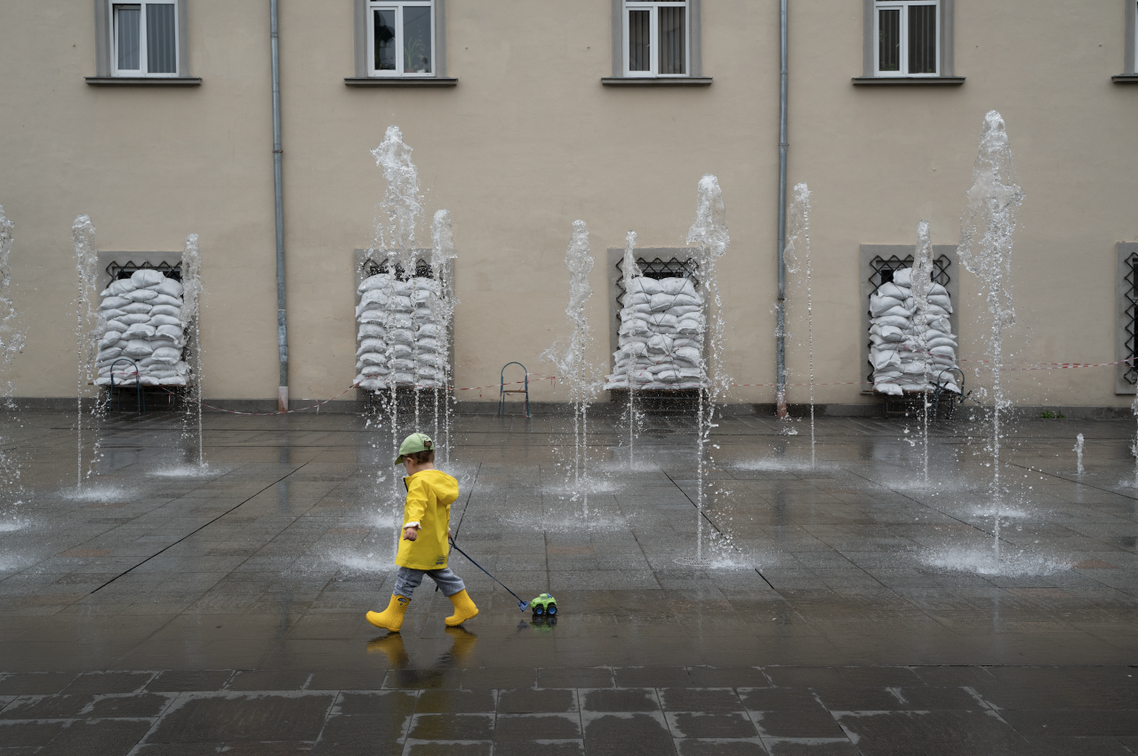 plac z fontanną, na pierwszym tle chłopiec ciągnący zabawkę, w tle budynek z oknami wyłożonymi workami z piaskiem